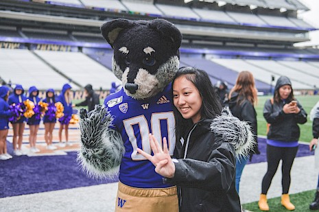 A student taking a picture with Harry the Husky on the Alaska Airlines Stadium Field holding up the "W" hand sign
